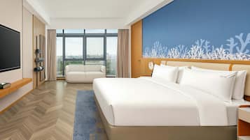 Habitación de hotel con una cama grande, televisión, zona de estar con sofá y vistas a la ciudad a través de las ventanas.