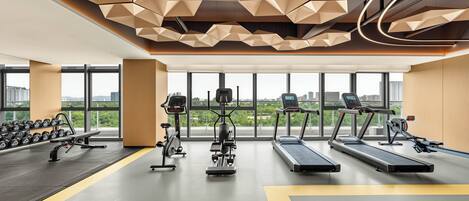 A modern gym with treadmills, elliptical machines, and dumbbells, large windows, and a geometric ceiling design.