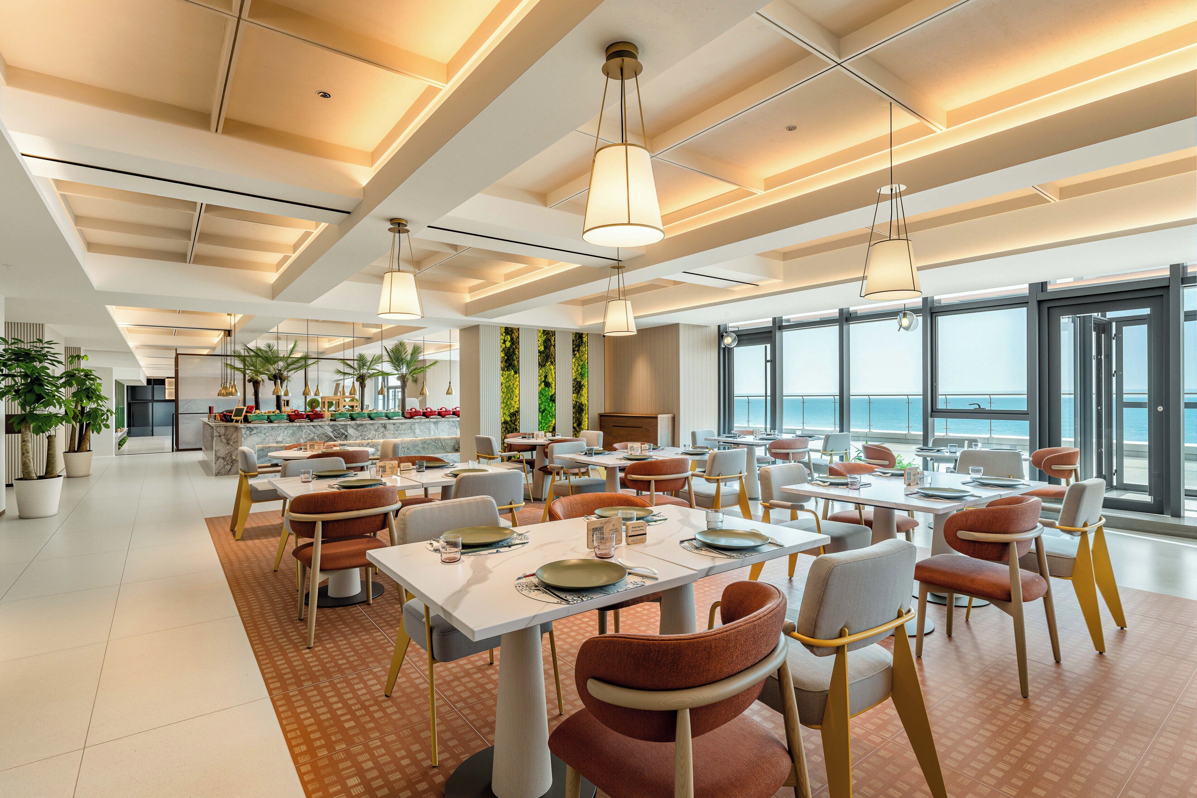 A modern dining area with tables and chairs, large windows, and a view of the sea.