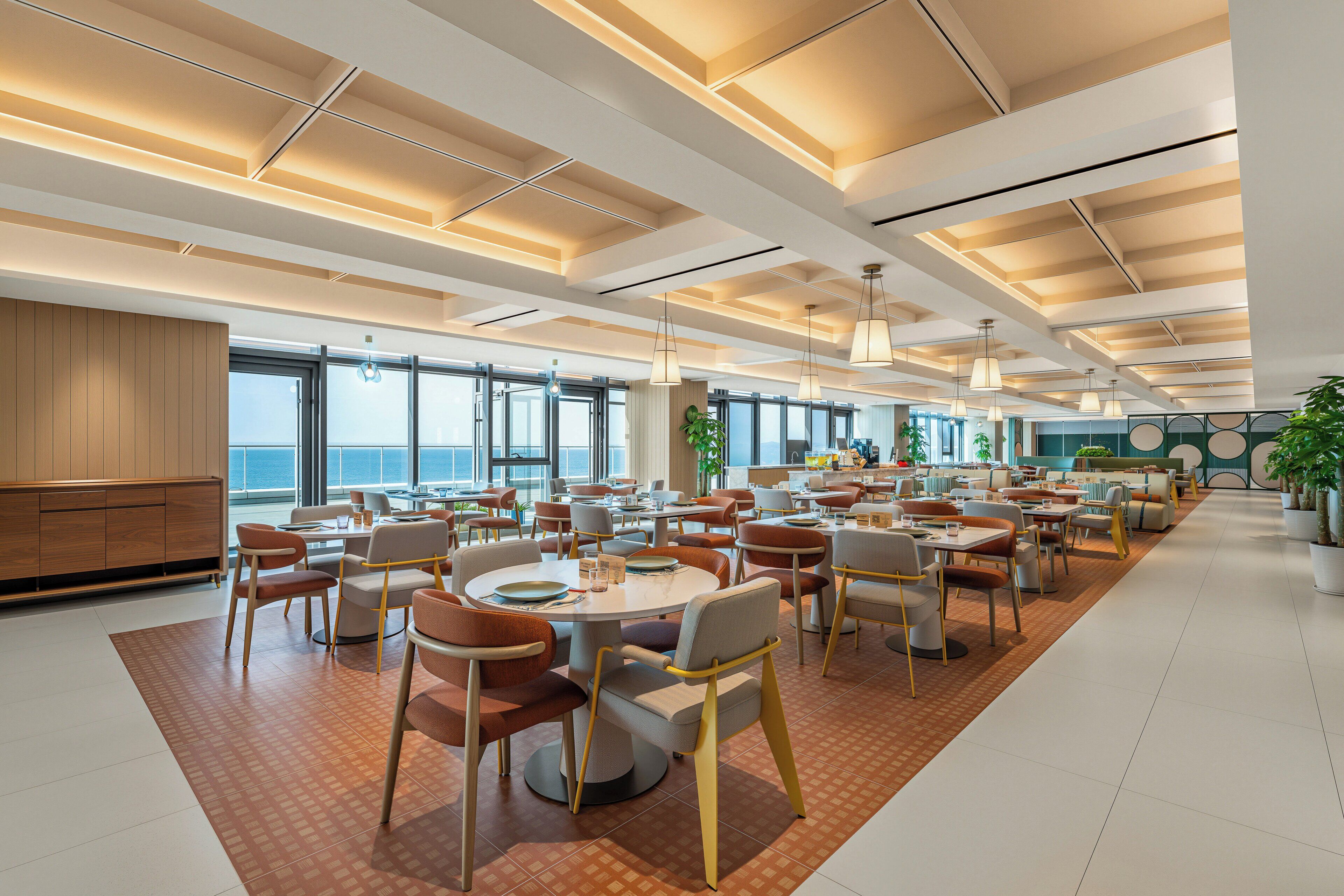 A spacious dining area with multiple tables and chairs, large windows offering a view of the sea, and a modern ceiling design.