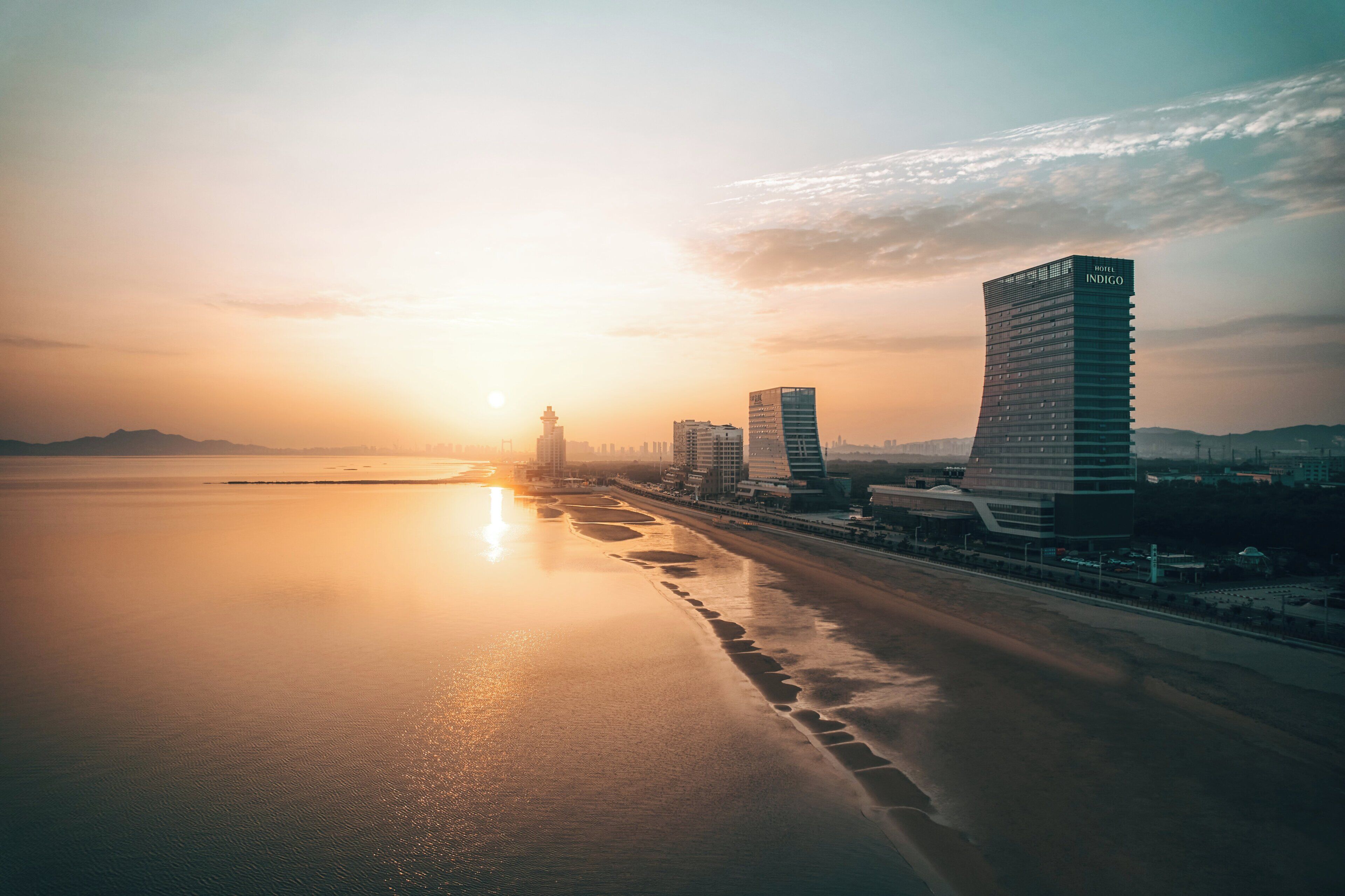 A coastal cityscape with modern buildings, including a hotel named 'Hotel Indigo,' during sunset.