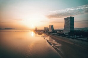 A coastal cityscape with modern buildings, including a hotel named 'Hotel Indigo,' during sunset.