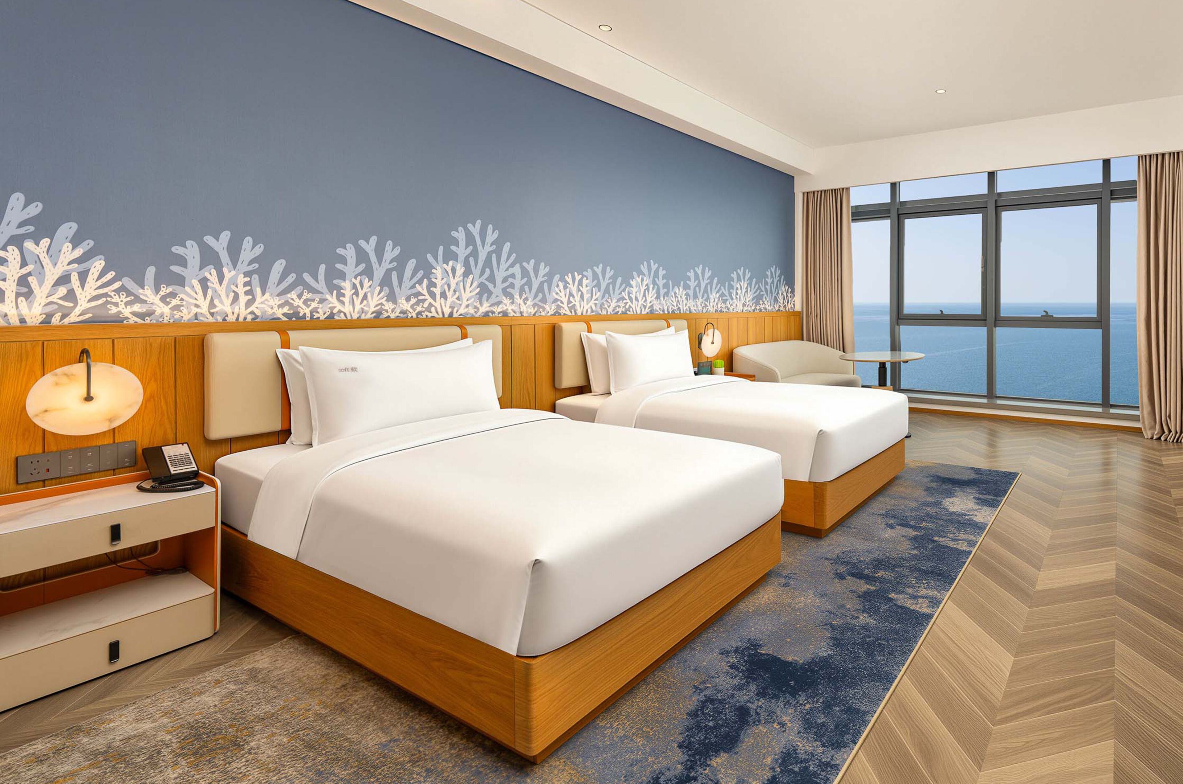 A hotel room with two beds, a desk, a lamp, and a view of the sea.