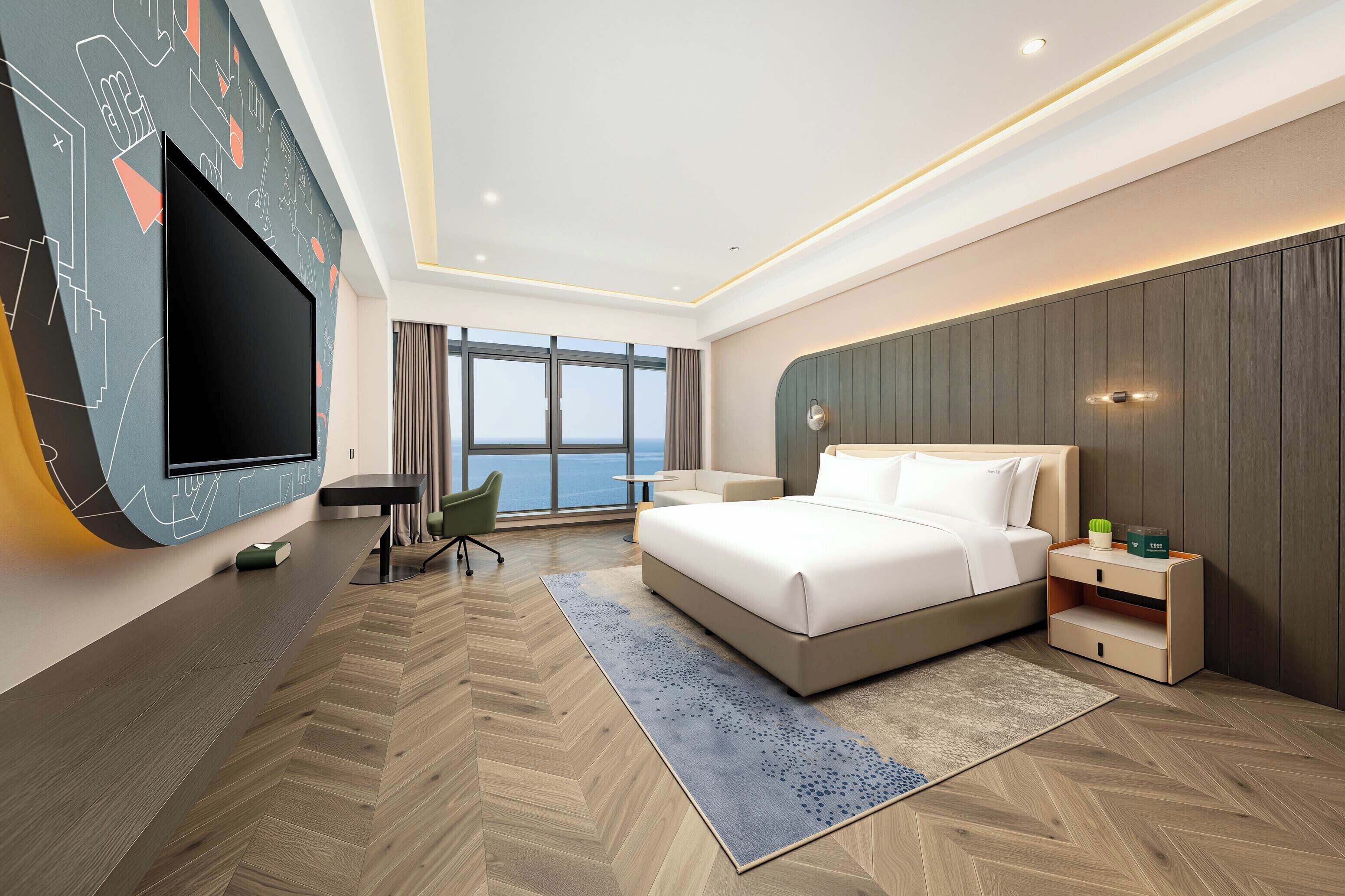 A modern hotel room with a large bed, a flat-screen TV mounted on the wall, a desk, and a view of the sea through large windows.