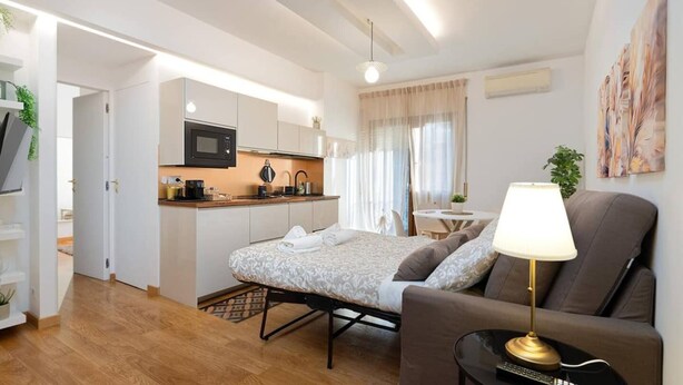 Habitación de hotel compacta con kitchenette, cama con colcha estampada, sofá con lámpara y un cuadro en la pared.