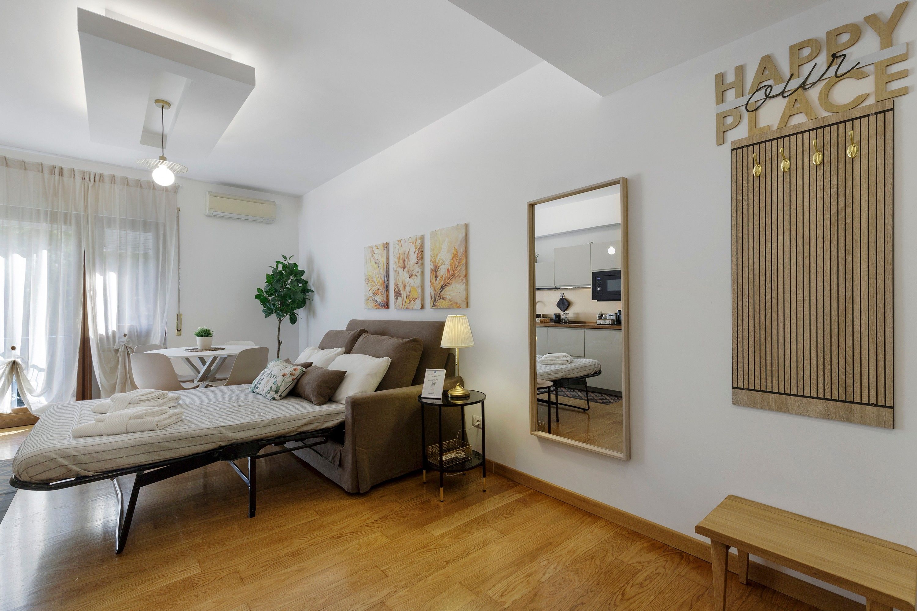 Una moderna camera d'hotel con un letto, un comodino, una lampada e uno specchio. È presente una zona pranzo con tavolo e sedie, e una pianta. La stanza ha il pavimento in legno e una luce a soffitto.