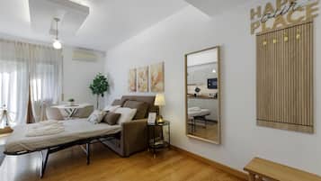 Una moderna camera d'hotel con un letto, un comodino, una lampada e uno specchio. È presente una zona pranzo con tavolo e sedie, e una pianta. La stanza ha il pavimento in legno e una luce a soffitto.