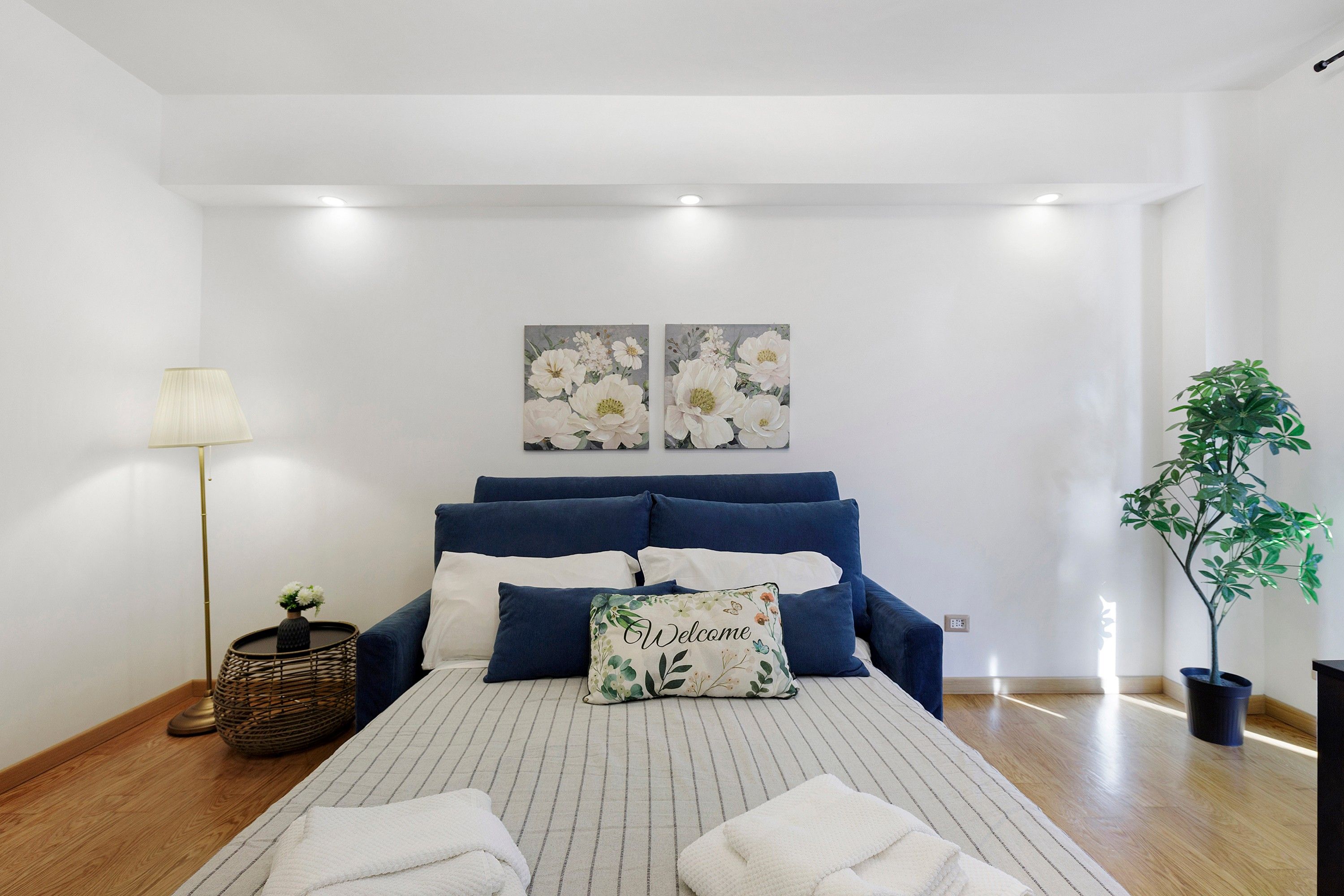 A bedroom with a bed, pillows, a bedside lamp, a plant, and artwork on the wall.