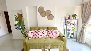 A living room with a green sofa, patterned cushions, a wall-mounted circular decoration, a plant, and a shelf with potted plants.