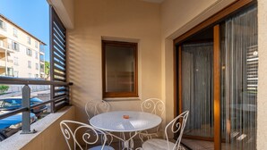 A balcony with a round table and four chairs, a window with a wooden frame, and a view of buildings and parked cars. - La Parenthèse d'Uzès - Avec Parking Privé (Uzes)