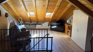 An attic room with wooden beams, a stone wall, a large TV, and a staircase. - Ca'Lina by Lago Maggiore Rentals & Estate (Cannobio)