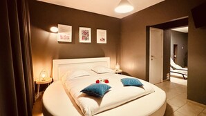 Uma cama redonda com lençóis brancos, duas almofadas, uma mesa de cabeceira com abajur e um quadro fixado na parede.
