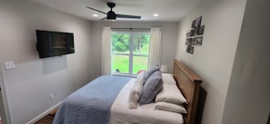 A bedroom with a bed, a ceiling fan, a TV mounted on the wall, and a window with curtains.