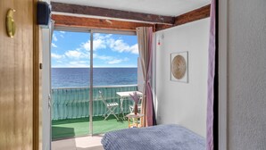 A room with a bed, a sliding glass door leading to a balcony with a view of the sea, a small table with a chair, and a fan on the wall.