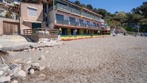 Ein mehrstöckiges Gebäude mit Balkonen, ein Strand im Vordergrund und eine hügelige Landschaft im Hintergrund.