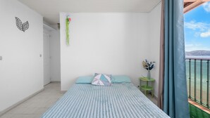 Un dormitorio con cama, una mesita de noche con un florero, una planta colgante y un balcón con vistas.