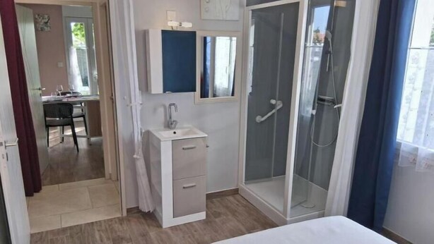 A bedroom with a bed, a walk-in shower, a small bathroom with a sink, and a dining area visible through an open door. - Charming apartment with swimming pool 6 km from the Vendée beaches (Notre-Dame-de-Riez)