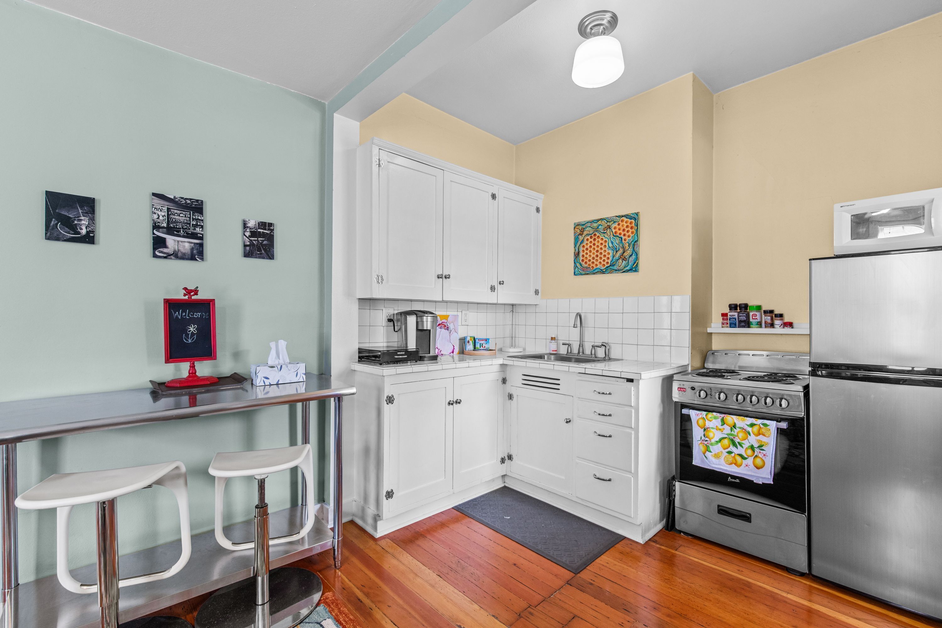 A kitchen with white cabinets, a stainless steel refrigerator, a stove, and a microwave. There are two bar stools and a welcome sign.
