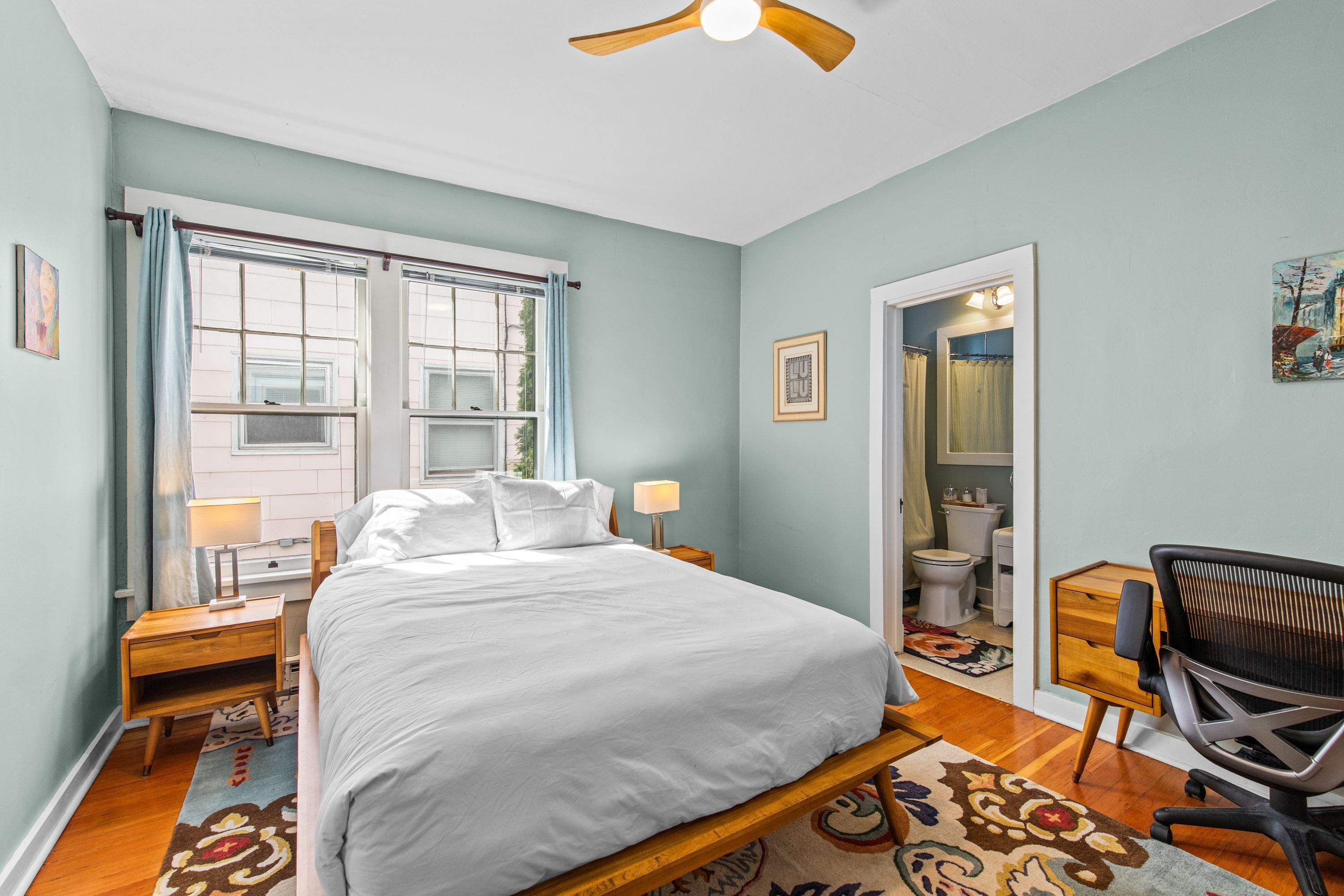 A bedroom with a bed, bedside tables, a desk, a chair, a ceiling fan, and a window with curtains.