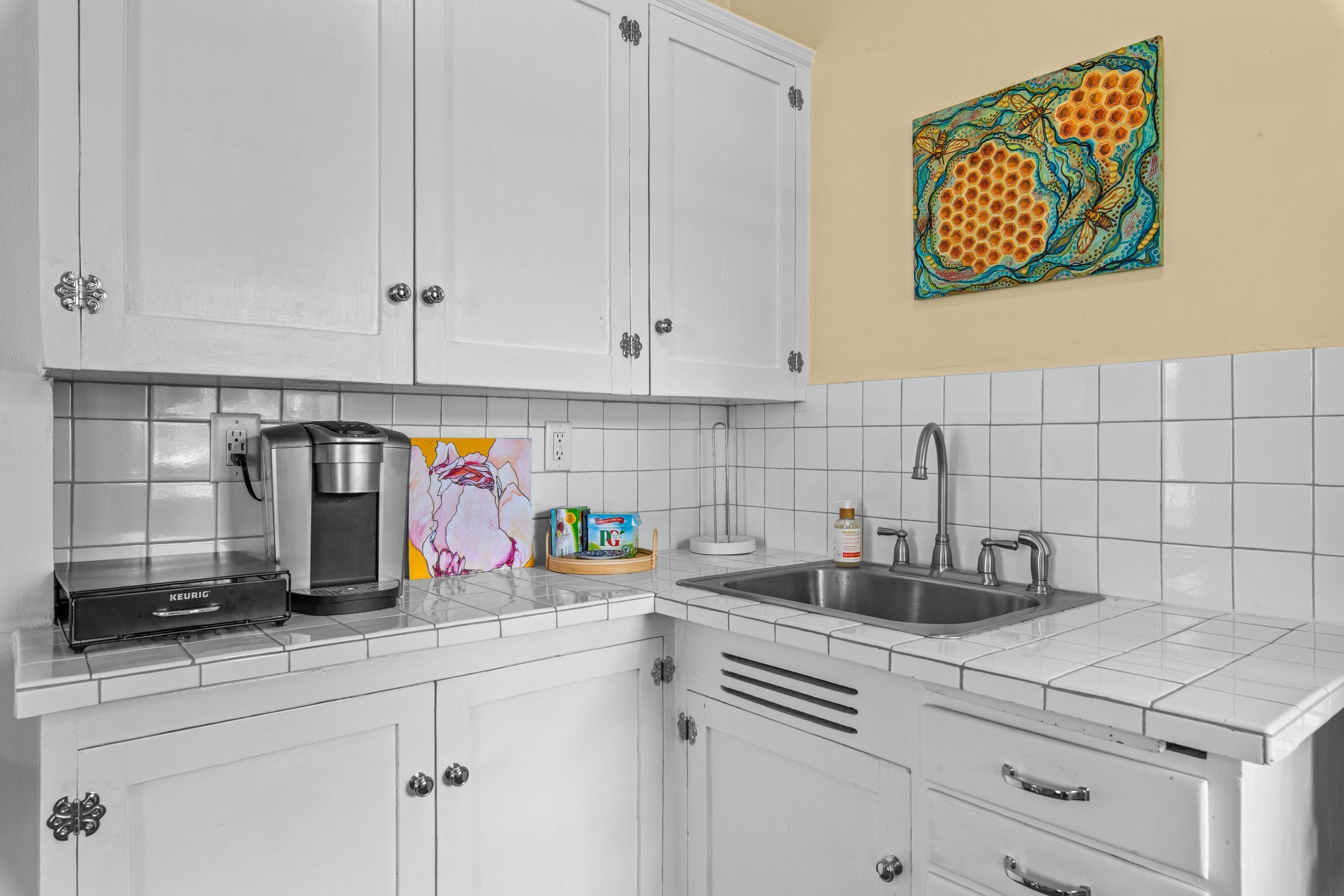 Una cocina con armarios blancos, una cafetera, un lavabo y un cuadro de colores en la pared.
