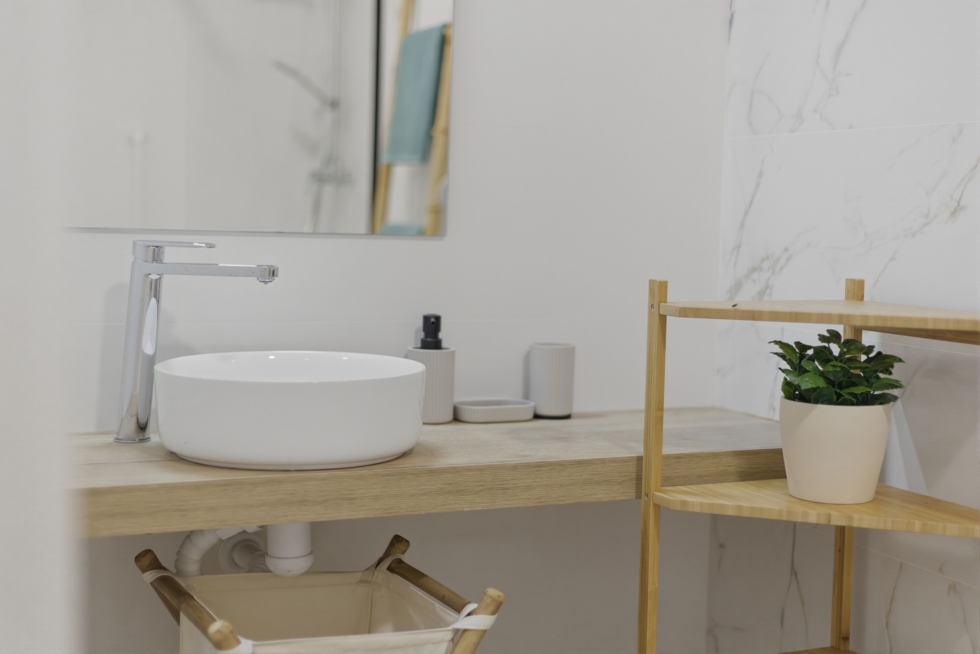 Un baño moderno con un lavamanos blanco redondo, un grifo cromado, una pequeña botella blanca y un estante de madera con una planta en maceta.