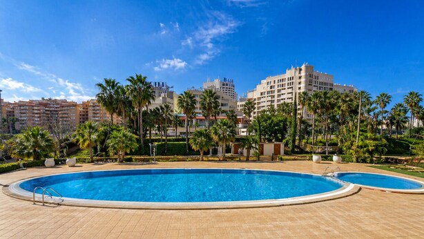 A swimming pool with a tiled deck, surrounded by palm trees and buildings in the background. - Spacious 2-bedroom Apartment With Pool View in Oropesa del Mar (Oropesa del Mar)