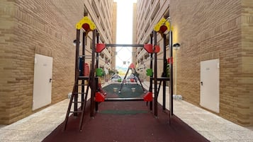 A playground structure with climbing and sliding elements, set against a brick building facade.