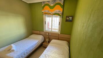 Een kamer met twee bedden, een raam met een gordijn met patroon, en een klein schilderij aan de muur.