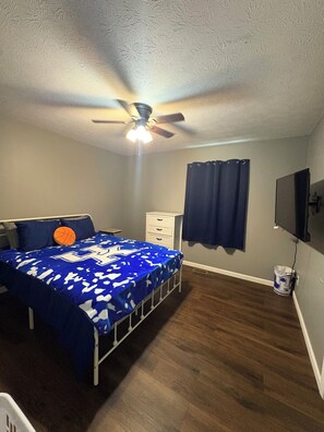 Un dormitorio con cama, ventilador de techo, cómoda, televisor y ventana con cortinas.