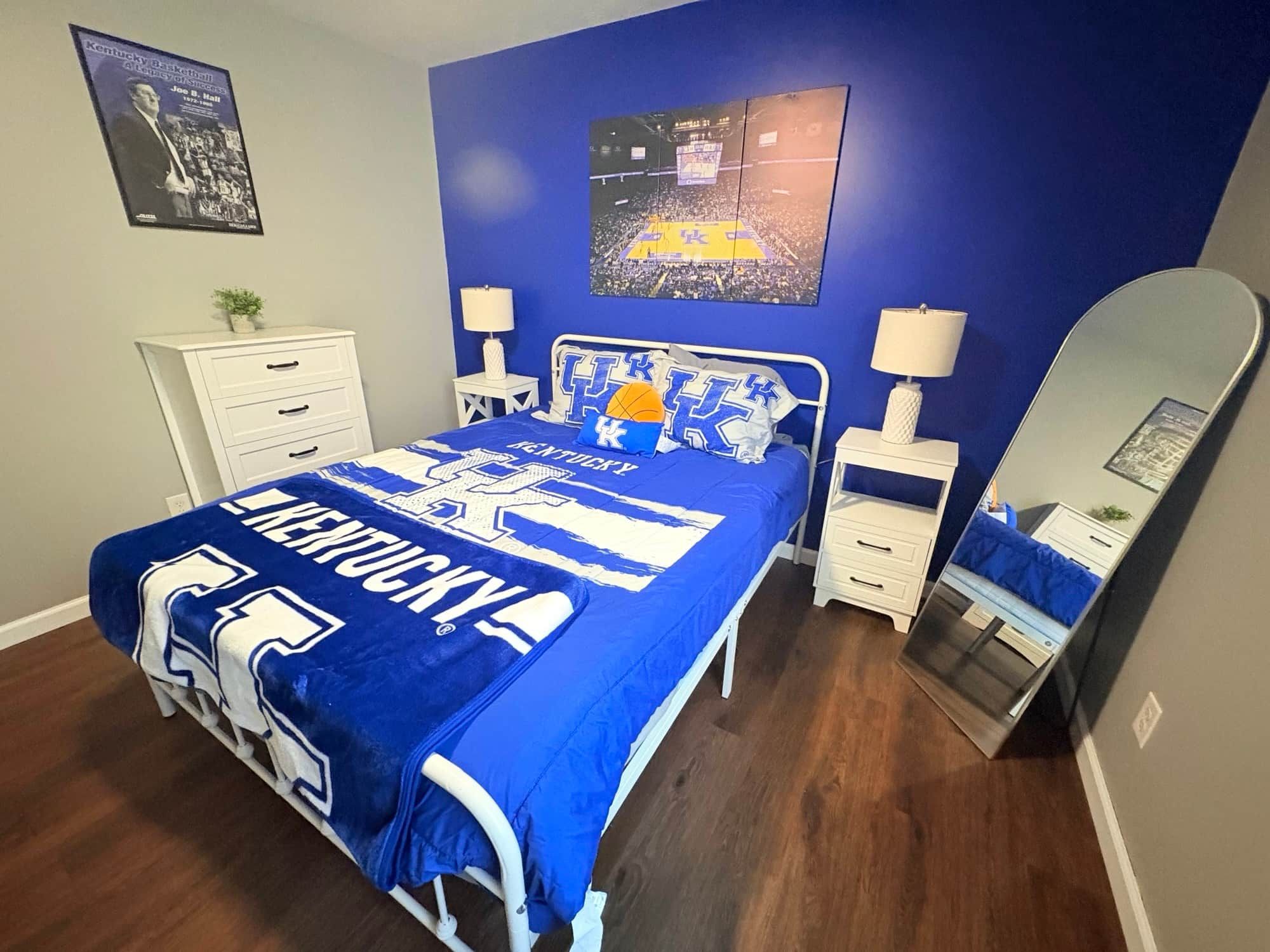 A bedroom with a bed featuring blue and white Kentucky University bedding, a white nightstand, a bedside table with a lamp, a mirror, and a framed picture on the wall.