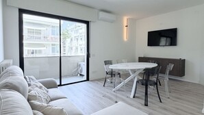Un soggiorno moderno con un divano, un tavolo da pranzo con sedie, una TV a parete e una porta vetrata scorrevole che dà su un balcone.