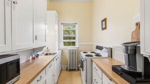 Una cucina compatta con mobili bianchi, un piano cottura, una macchina per il caffè e una finestra vista alberi.