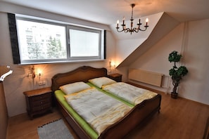 Ein Schlafzimmer mit Bett, Nachttischen, einem Fenster mit Jalousien, einer Heizung, einer Pendelleuchte und einem Blumentopf.