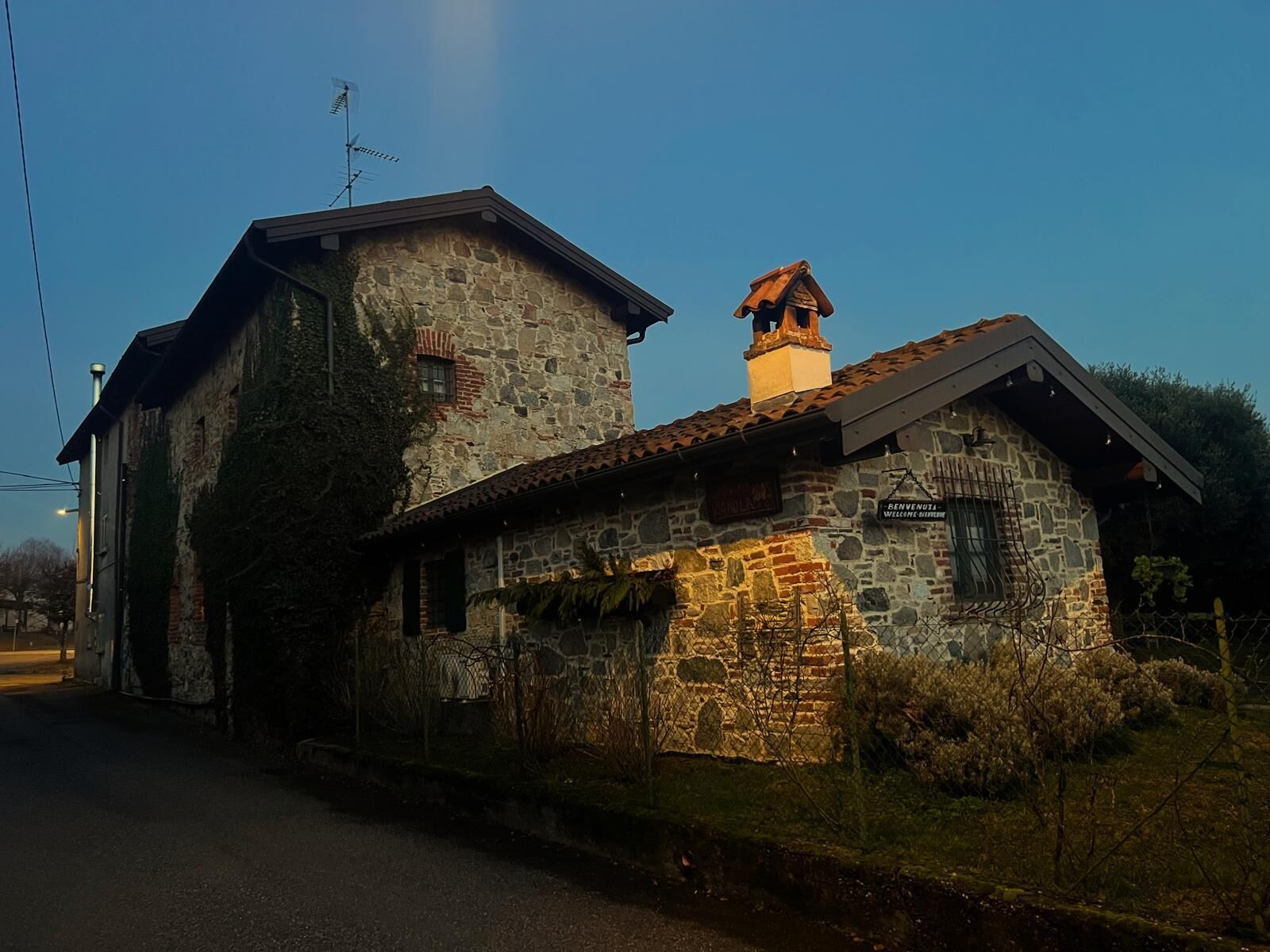 Ein Steinhaus mit Schornstein, einem Schild mit der Aufschrift 'Benvenuti Welcome' und einem kleinen Fenster.