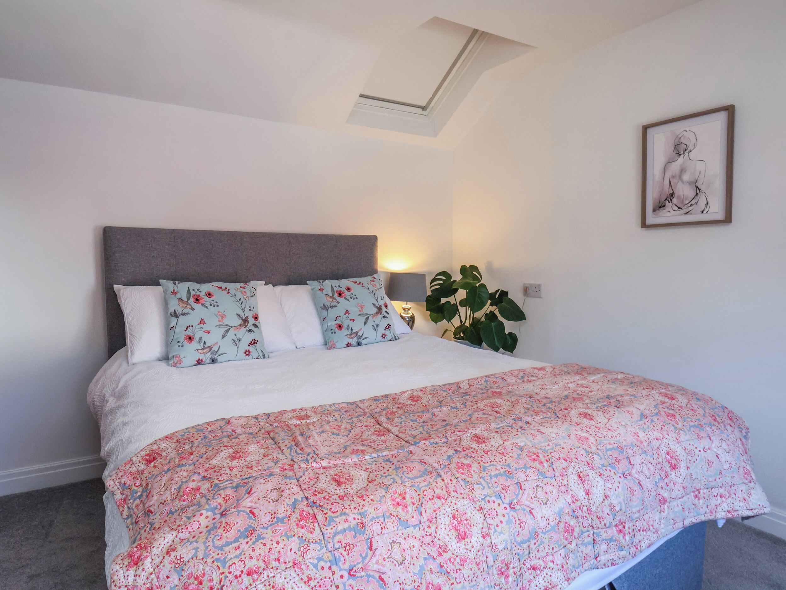 A bedroom with a bed, floral bedding, a grey headboard, a skylight, a potted plant, and a framed artwork on the wall.