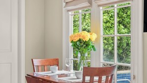 Une table à manger avec des chaises, un vase rempli de fleurs jaunes et une fenêtre donnant sur un paysage verdoyant et une piscine.