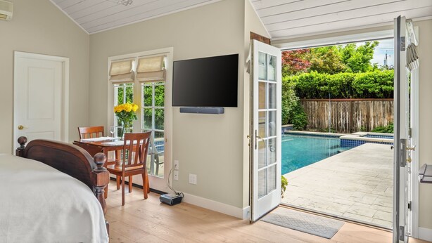 Une chambre avec un lit, un téléviseur fixé au mur, une table à manger avec des chaises, et une vue sur une piscine par une porte ouverte.