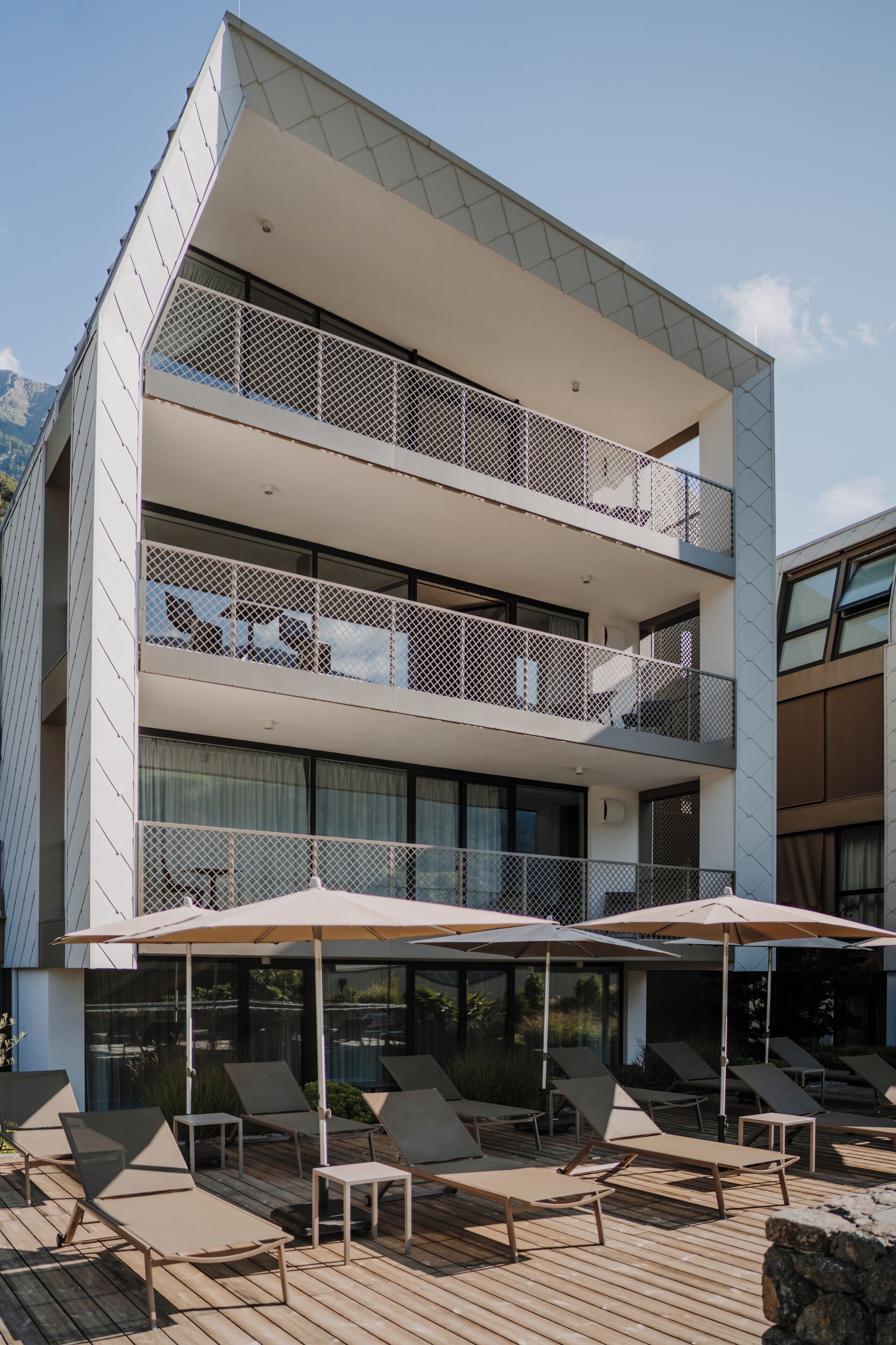 Um edifício moderno de vários andares com um terraço no telhado, com cadeiras de descanso e guarda-sóis.
