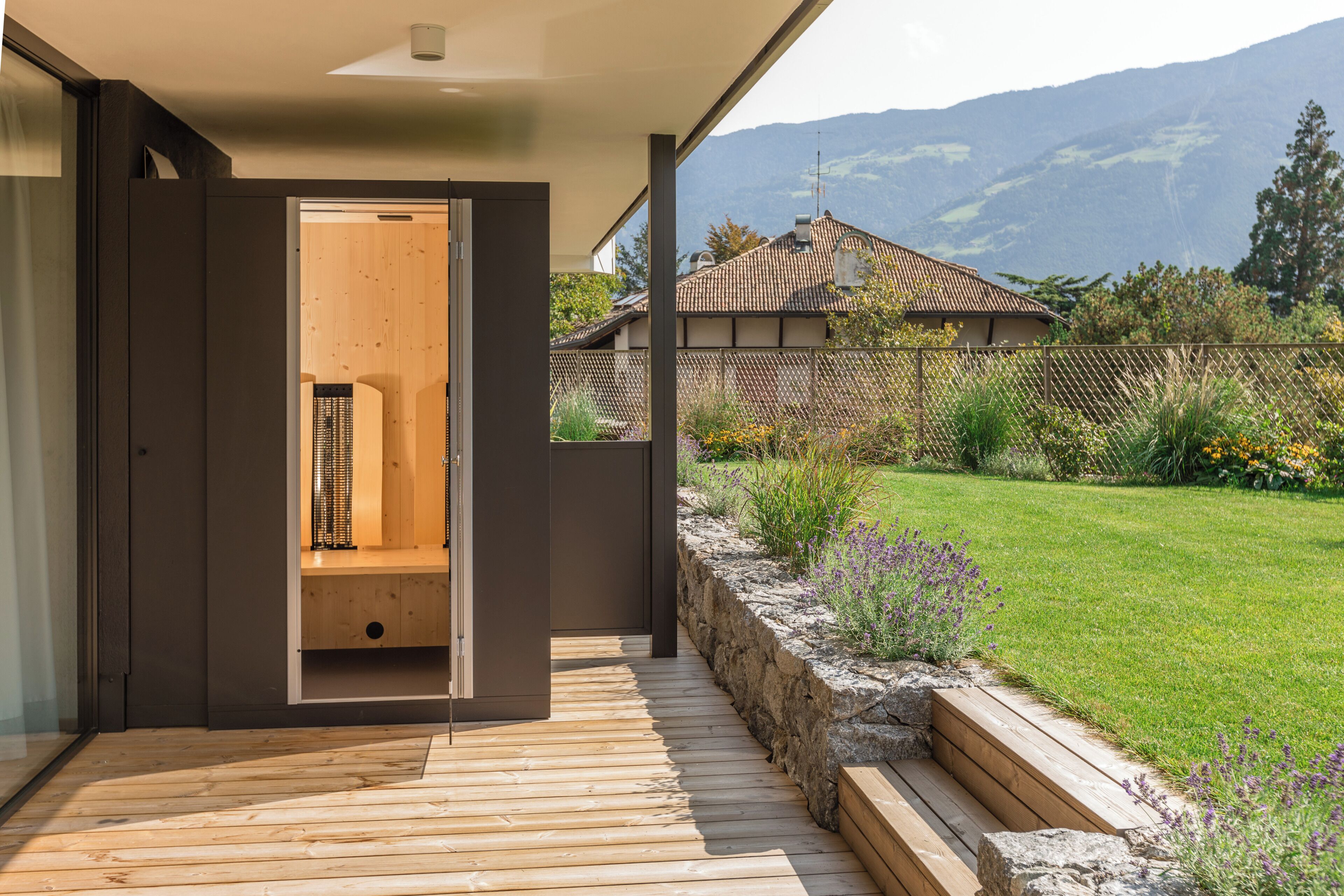 Une terrasse en bois mène à une pièce dotée d’une porte coulissante, un mur de soutènement en pierre et une vue sur une maison et des montagnes en arrière-plan.
