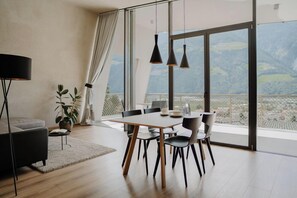 A modern dining area with a table and chairs, a sofa, a lamp, and a view of a mountainous landscape through large windows. - Marchegg Apartments (Naturno)