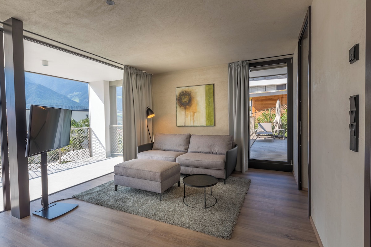 A modern living room with a sofa, ottoman, and TV, a painting on the wall, and a view of the outdoors.