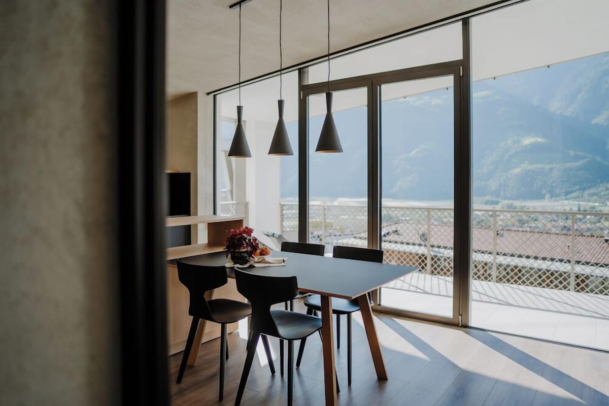 A modern dining area with a large table and chairs, pendant lights, and a view of the city through large windows.