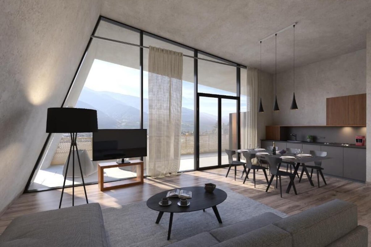 A modern living room with a sofa, a coffee table, a TV on a stand, a dining area with chairs, a kitchen with cabinets, and large windows offering a view.