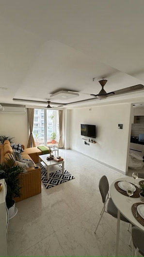A modern living room with a sofa, coffee table, dining table set for two, a TV mounted on the wall, and a ceiling fan.