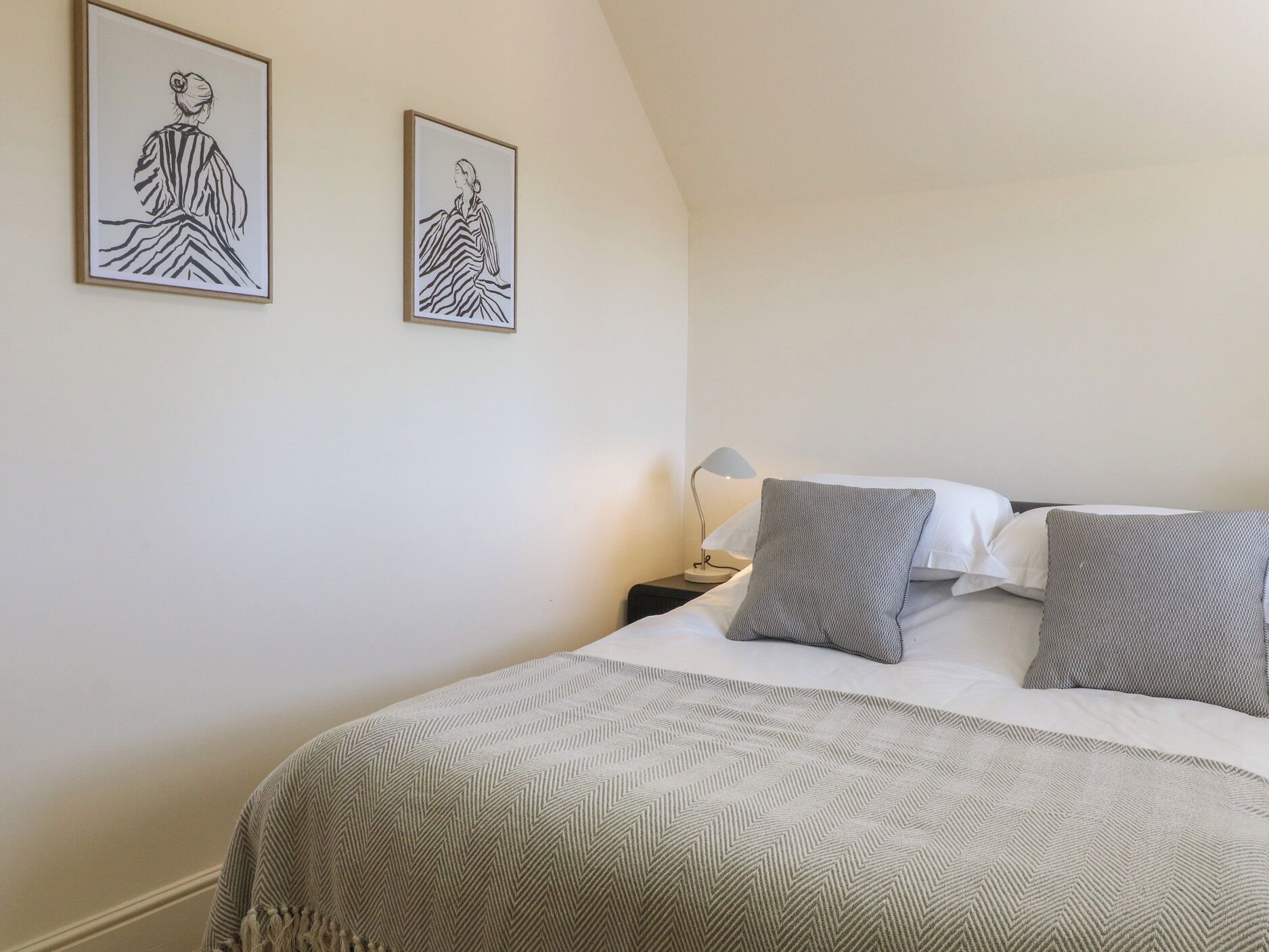 A bedroom with a bed, pillows, a nightstand, a lamp, and two framed artworks on the wall.