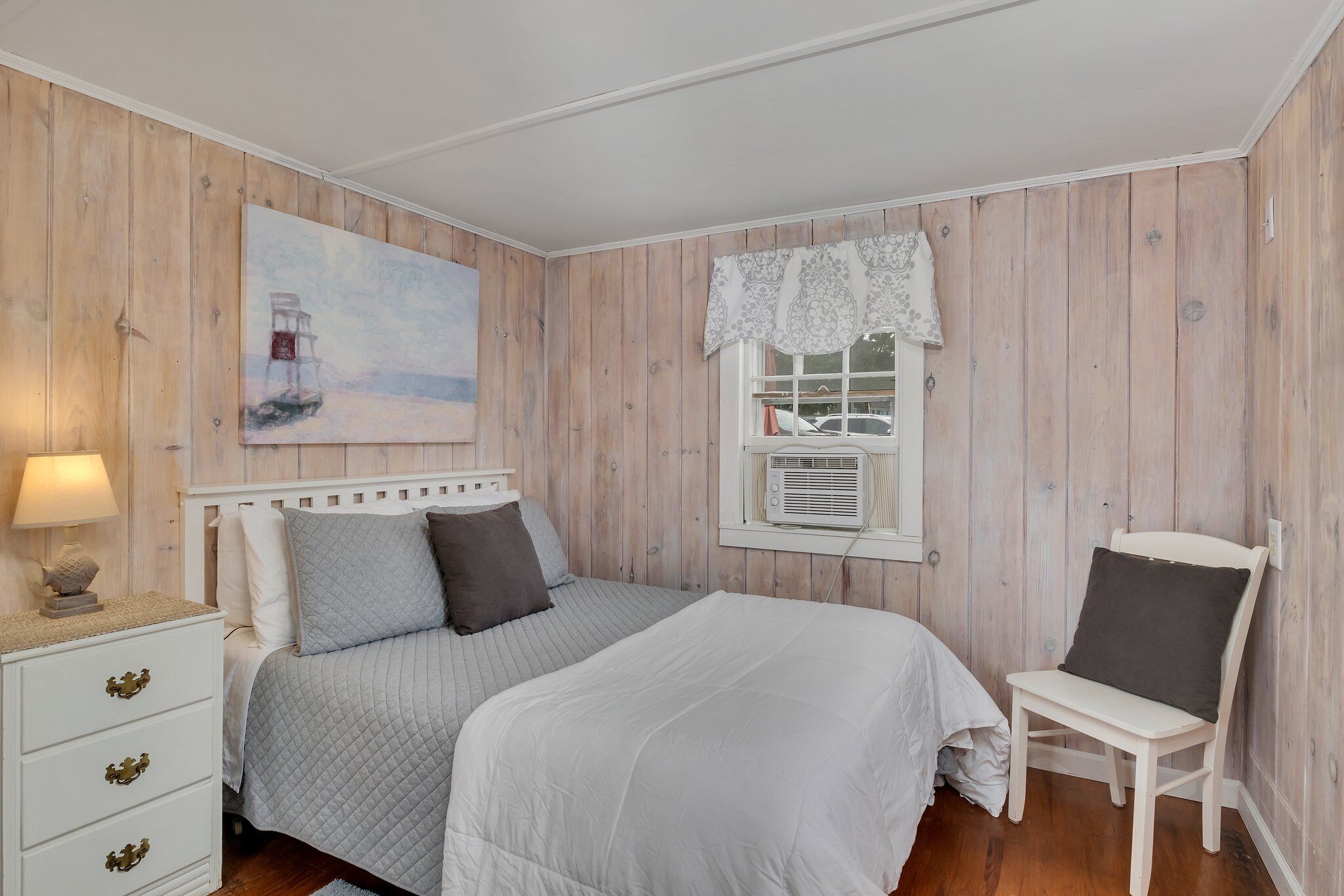Ein Schlafzimmer mit Bett, Nachttisch, Stuhl, Fenster mit Vorhang und einem Bild an der Wand.