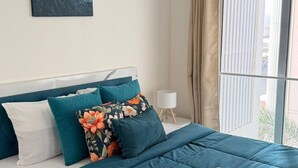 A bedroom with a bed, a nightstand, a lamp, a window with curtains, an air conditioning unit, and a painting on the wall. - Earthen Escape 2BHK near BKC Jio  Gym Access (Mumbai)