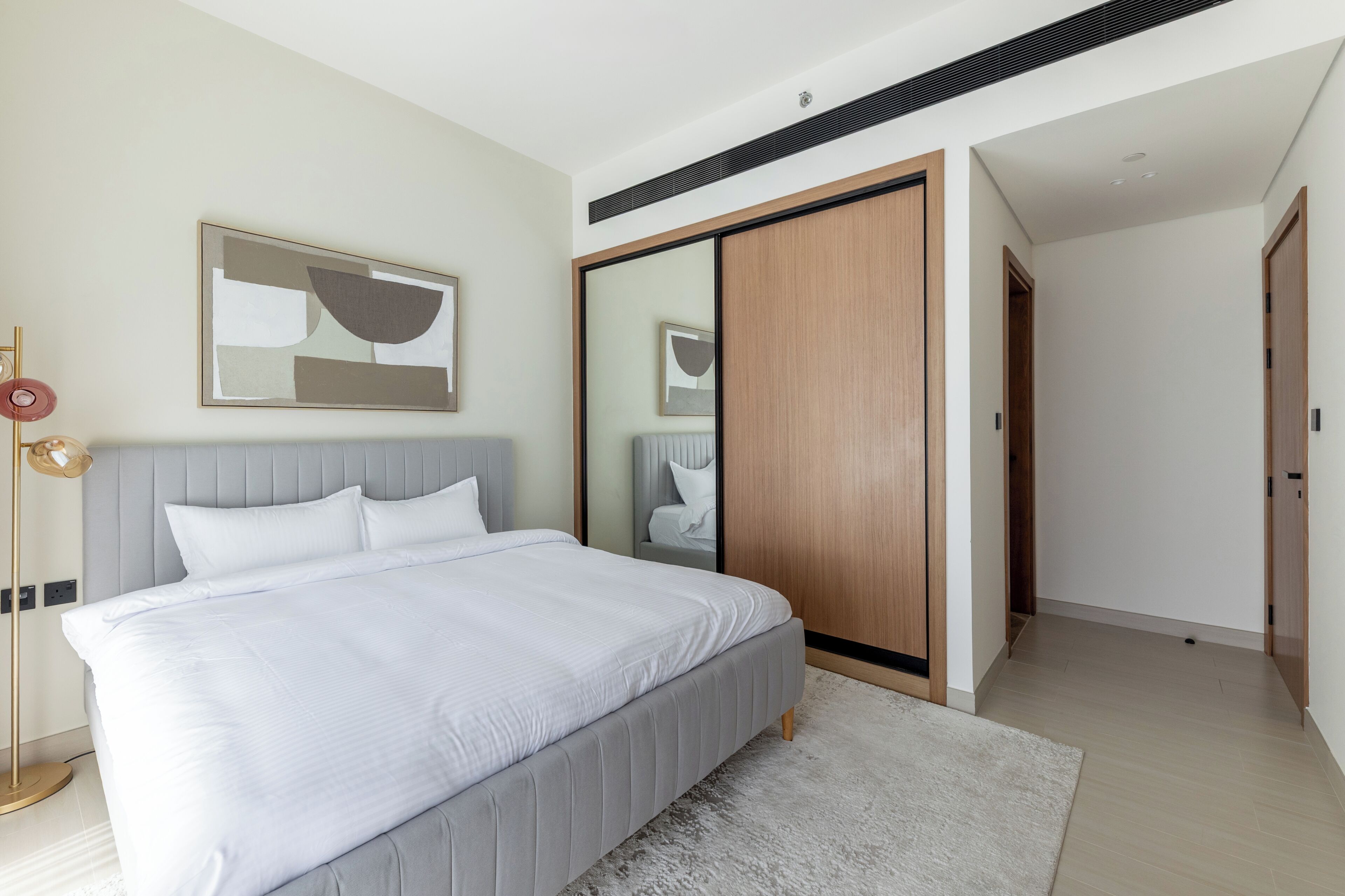 A modern bedroom with a large bed, a wardrobe, a bedside lamp, and a framed abstract painting on the wall.