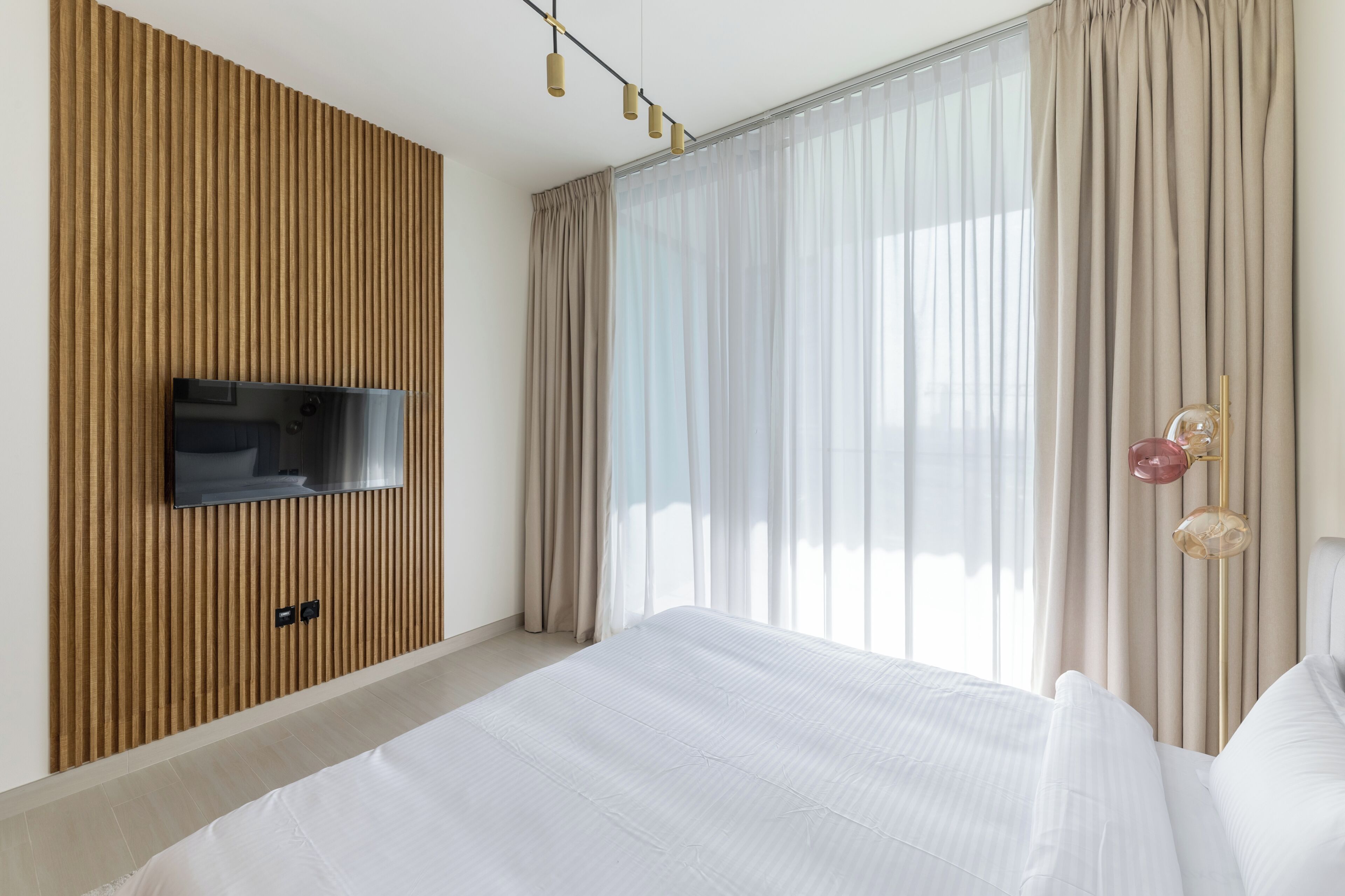 A modern hotel room with a large bed, a flat-screen TV mounted on a wooden panel wall, and a large window with sheer curtains.