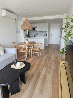 A modern living room with a sofa, dining table, and chairs, a kitchen in the background, and a wooden floor. - Urban 1303 | 2 rooms with garage (Mar del Plata)
